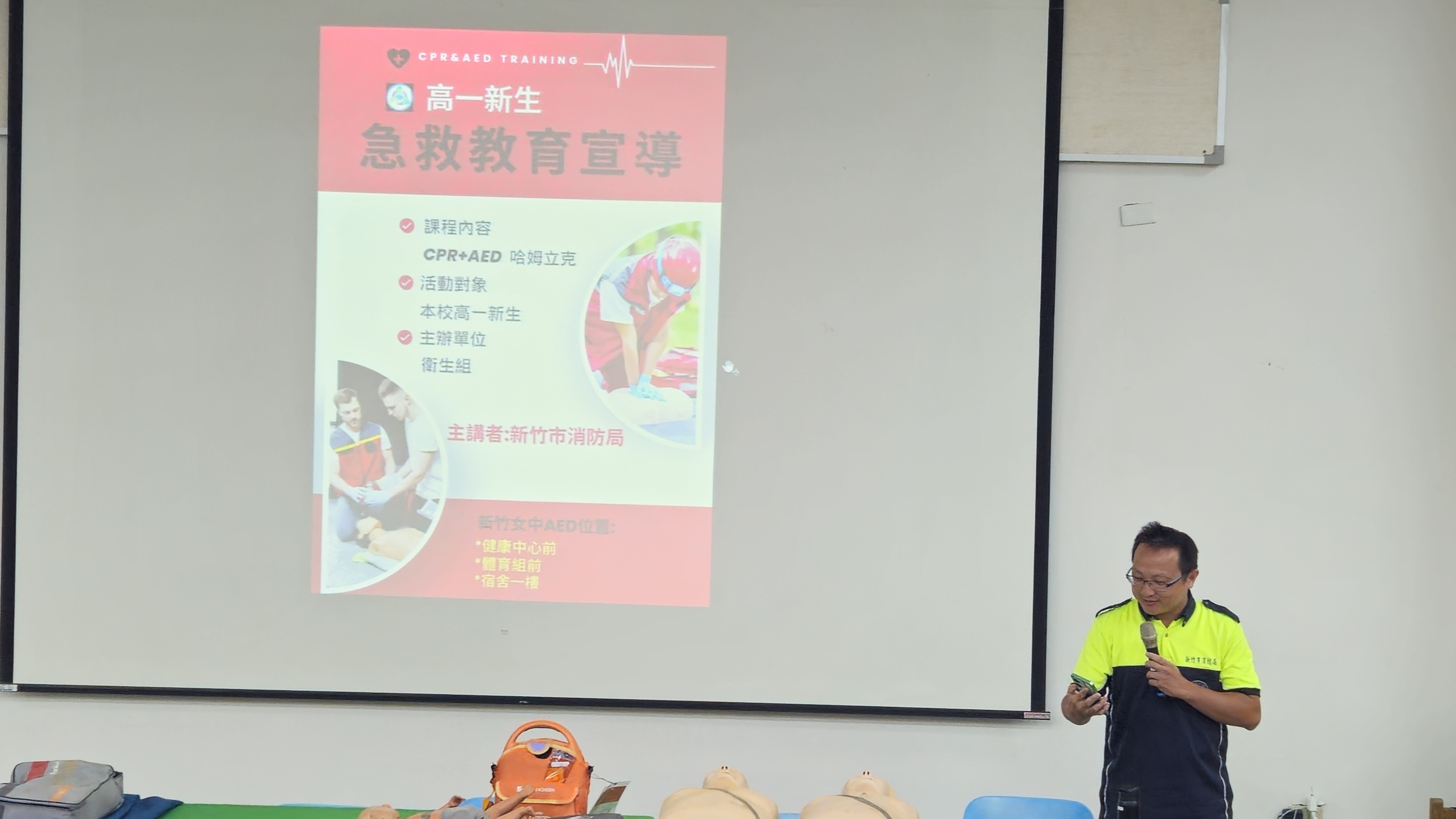 新竹市消防局講師於講台前介紹高一新生急救教育宣導，投影幕顯示簡報內容，前方擺放急救訓練器材。