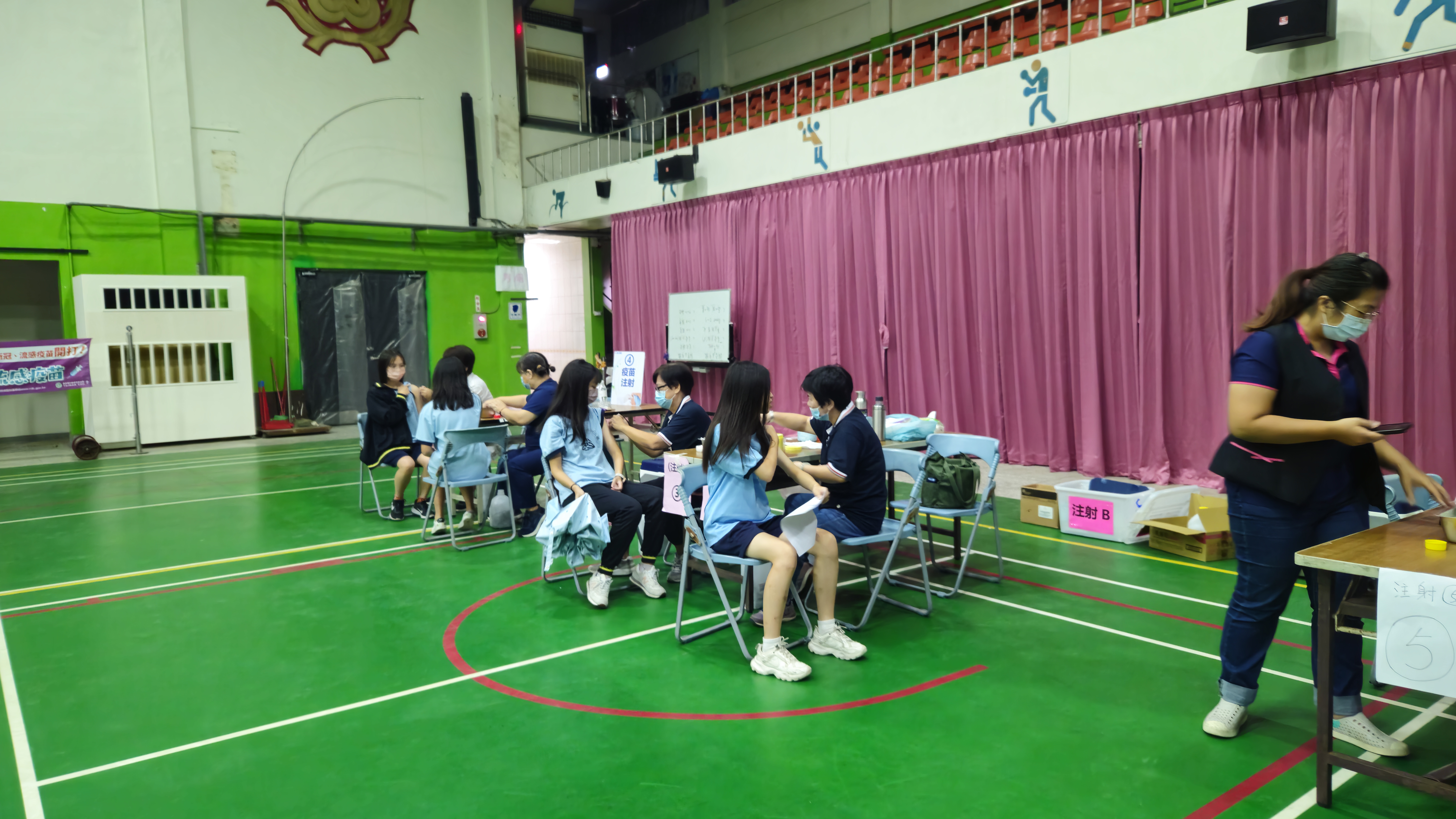 學生們坐在體育館一角排好的椅子上，由數名醫護人員同時進行流感疫苗注射。